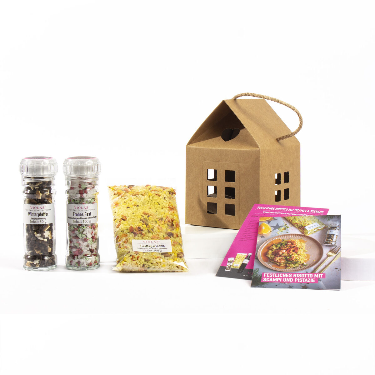 Ein Geschenkset bestehend aus einem Haus aus braunem Papier mit ausgeschnittenen Fenstern, zwei Gewürzmühlen, einer durchsichtigen Verpackung mit gelb-rotem Inhalt und zwei Rezeptkarten auf weißem Hintergrund.