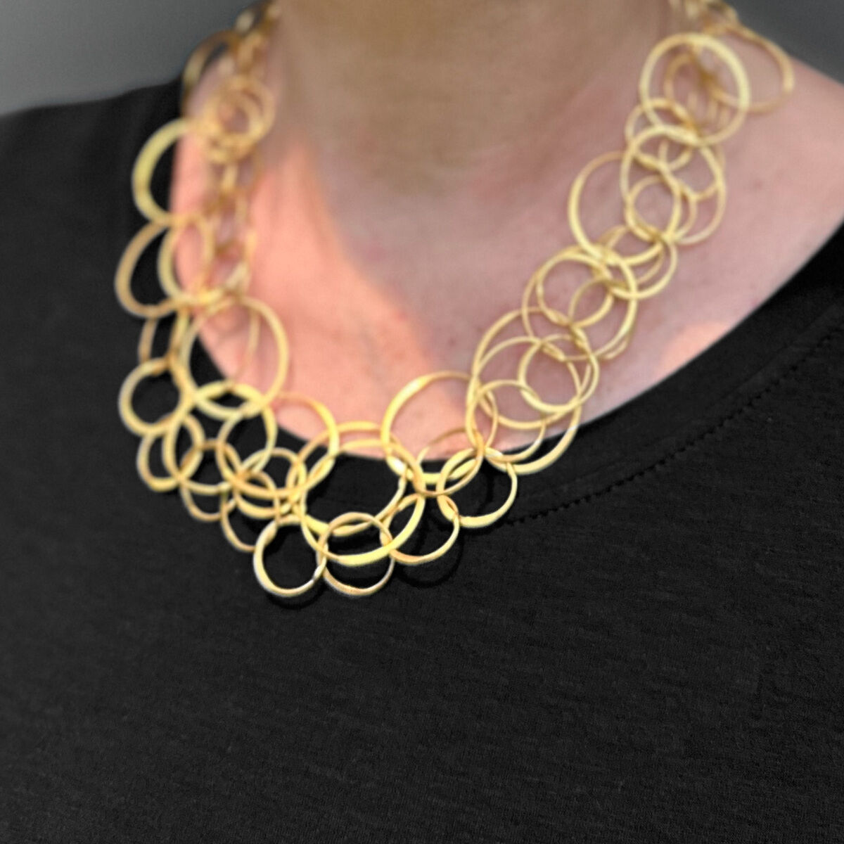 Eine Frau mit blonden Haaren und Brille trägt eine Halskette aus vergoldetem Silber, die aus ineinandergreifenden Ringen besteht. Sie trägt ein schwarzes Oberteil und hat einen neutralen Gesichtsausdruck.