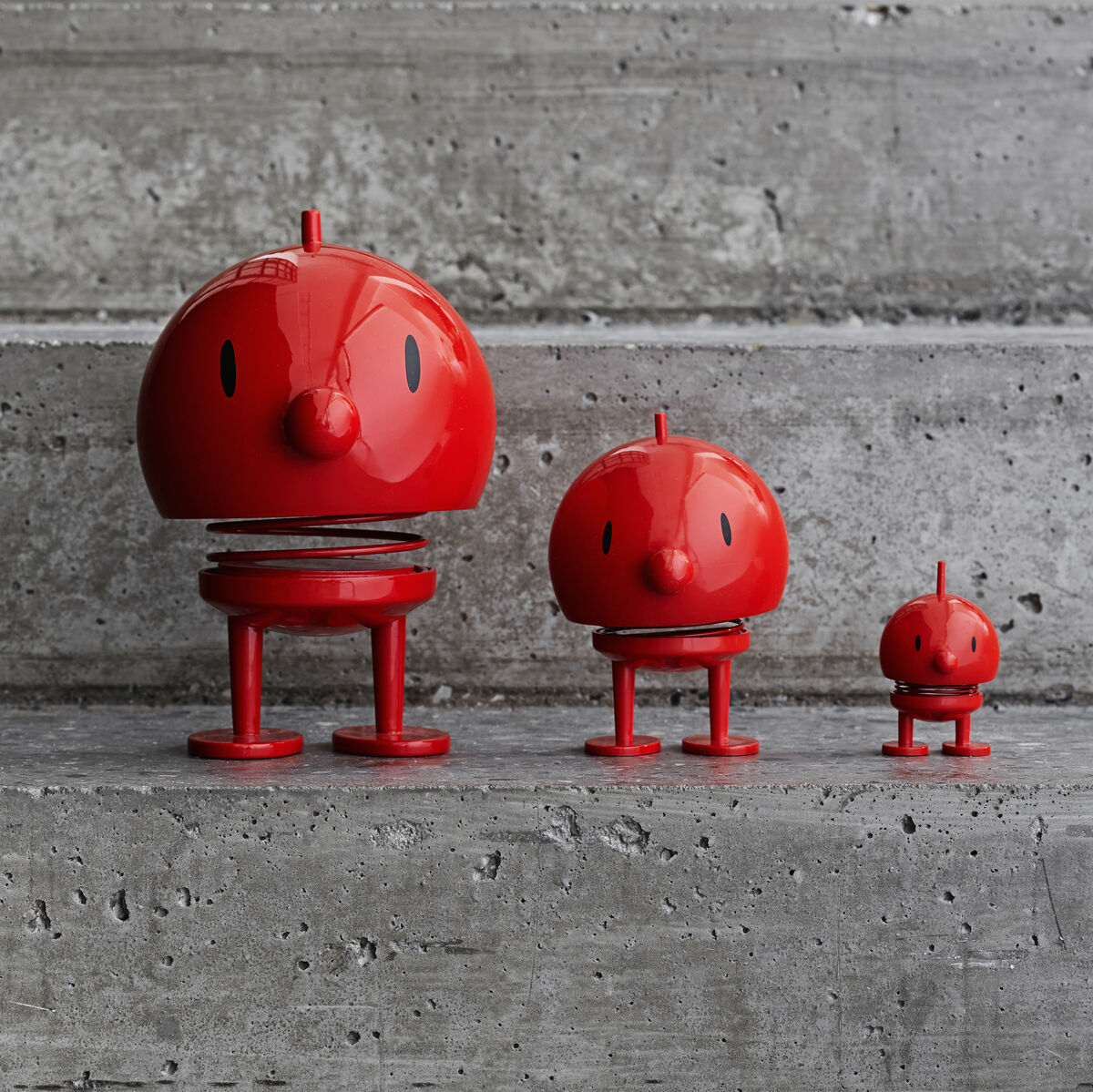 Drei rote Hoptimist Bumble Figuren in abnehmender Größe auf grauem Hintergrund. Die Figuren haben eine runde Form und lächeln fröhlich.