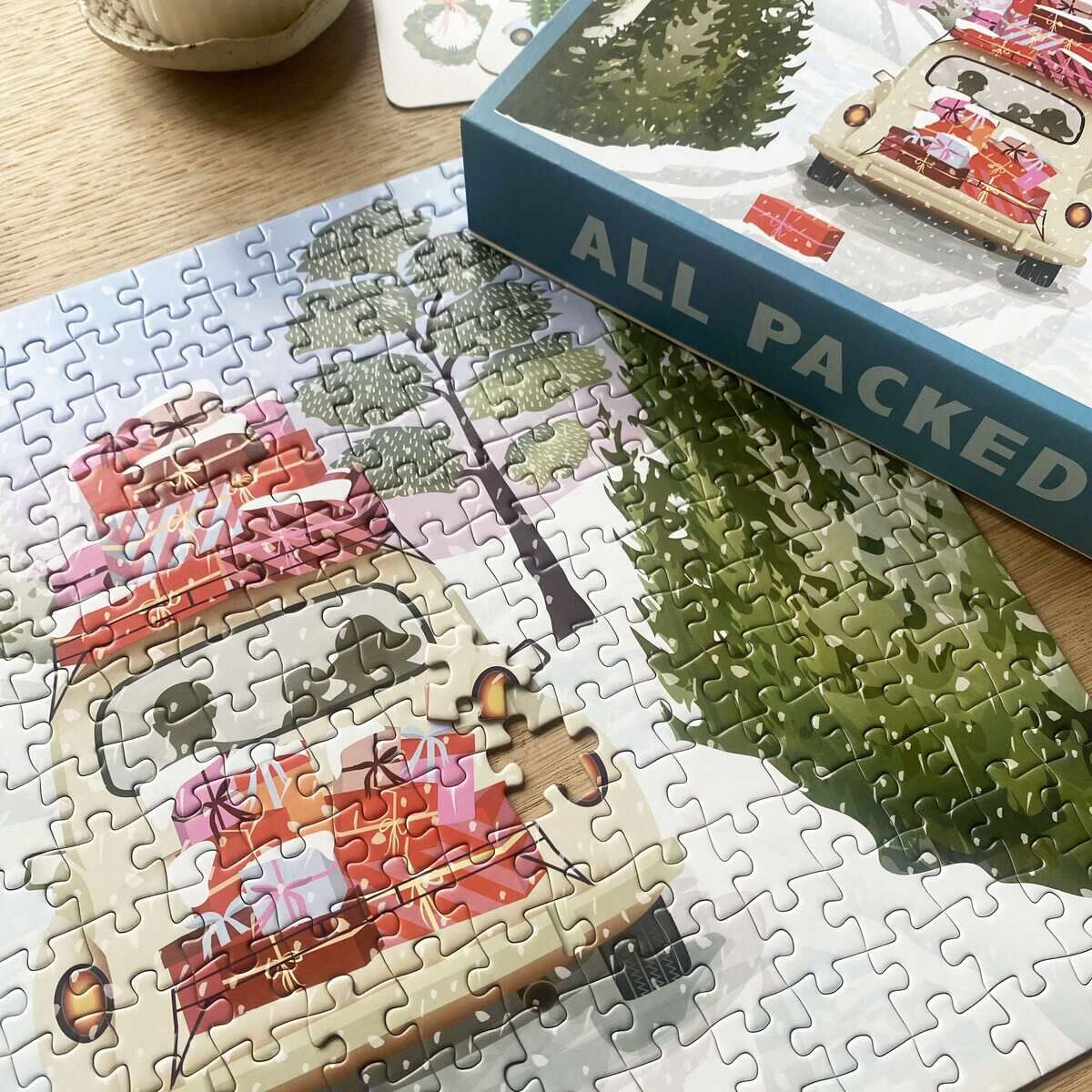 Das Bild zeigt das ViSSEVASSE 300-Teile-Puzzle »Alles gepackt«, fast vollständig zusammengesetzt. Es zeigt ein Auto voller bunter Geschenke auf schneebedeckter Straße im Wald. Die Verpackung liegt daneben. Ein letzter Puzzlestein fehlt in der Fahrzeugfront.