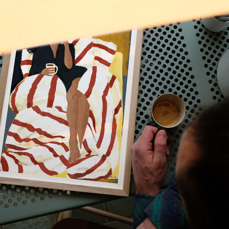 Gerahmtes Poster zeigt eine stilisierte Illustration einer Person mit einem Kaffee und einem Buch, eingerahmt von gestreifter Bettwäsche, fotografiert auf einem metallenen Tisch mit einer echten Tasse Kaffee daneben. Die Szene vermittelt Gemütlichkeit und Lesefreude.