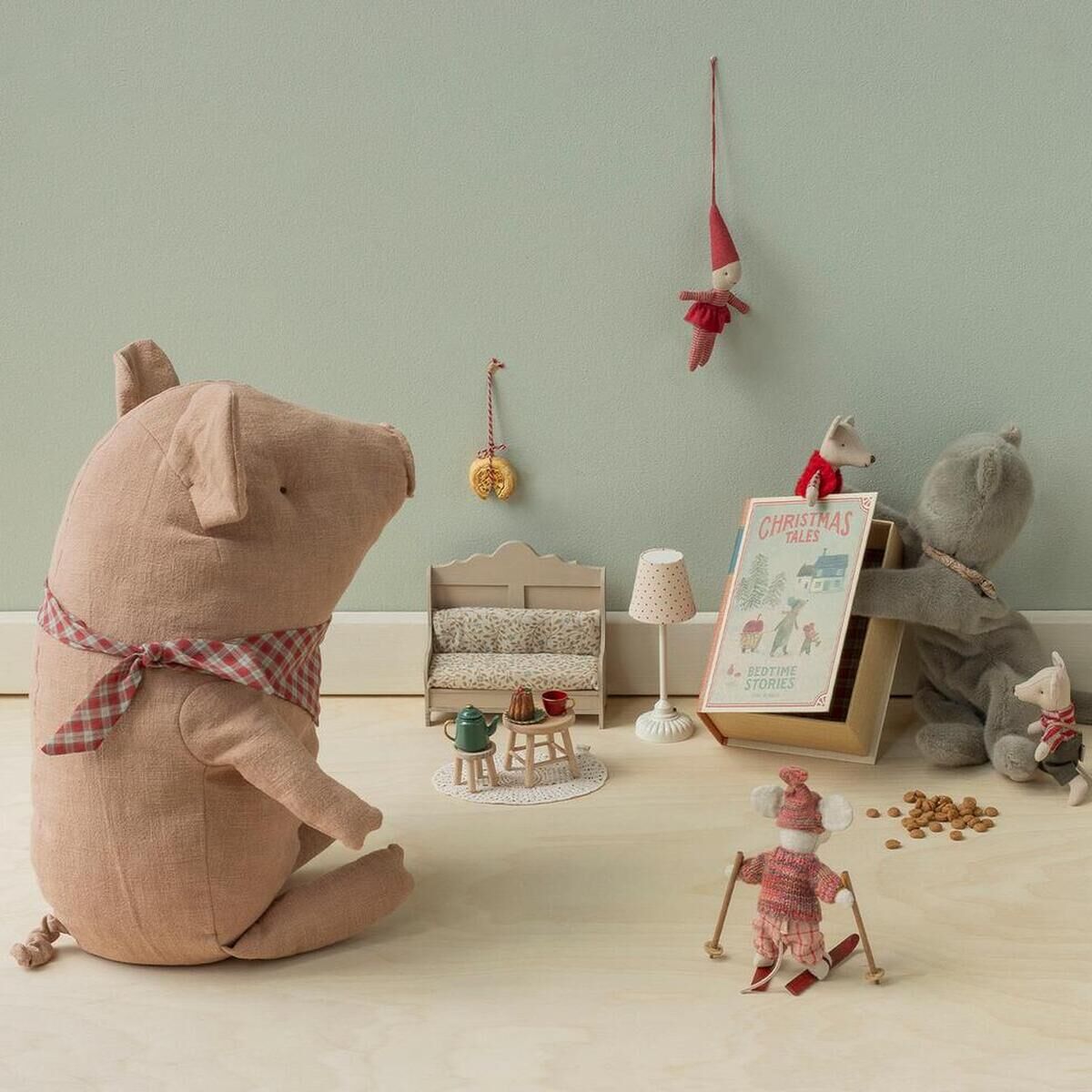 Das Bild zeigt das Maileg Kuscheltier »Großes Trüffelschwein« sitzend mit kariertem Schal in beige-rosafarbenem Stoff in einem kindlich dekorierten Raum. Im Hintergrund sind kleine Stofffiguren, Miniaturmöbel und Deko, alles in sanften Pastelltönen, arrangiert.