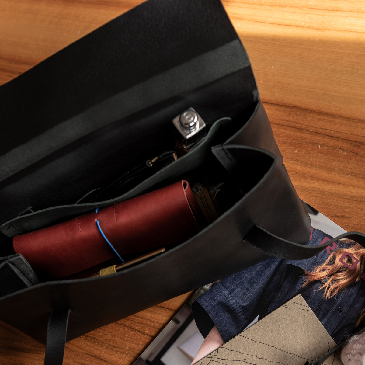 Eine schwarze Schultertasche aus Sattlerleder von Harold's Lederwaren liegt geöffnet auf einem Holztisch. Im Inneren befinden sich ein rotes Lederetui und weitere persönliche Gegenstände. Daneben liegen eine Brille, Hefte und ein Stein auf Zeitschriften mit Modemotiven.