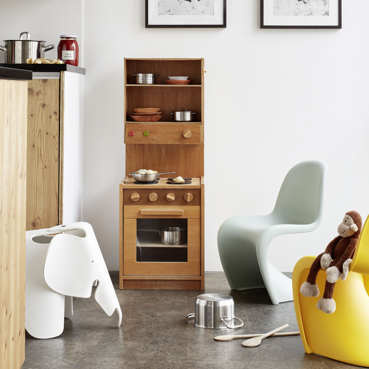 Ein moderner Kinderraum mit einem weißen Eames Elephant Hocker, einem gelben Kinderstuhl, einem Holzhocker mit Spielküche und einer plüschigen Spielzeugaffe. Der Raum hat zwei schwarz-weiße Fotos an der Wand und viel natürliches Licht.