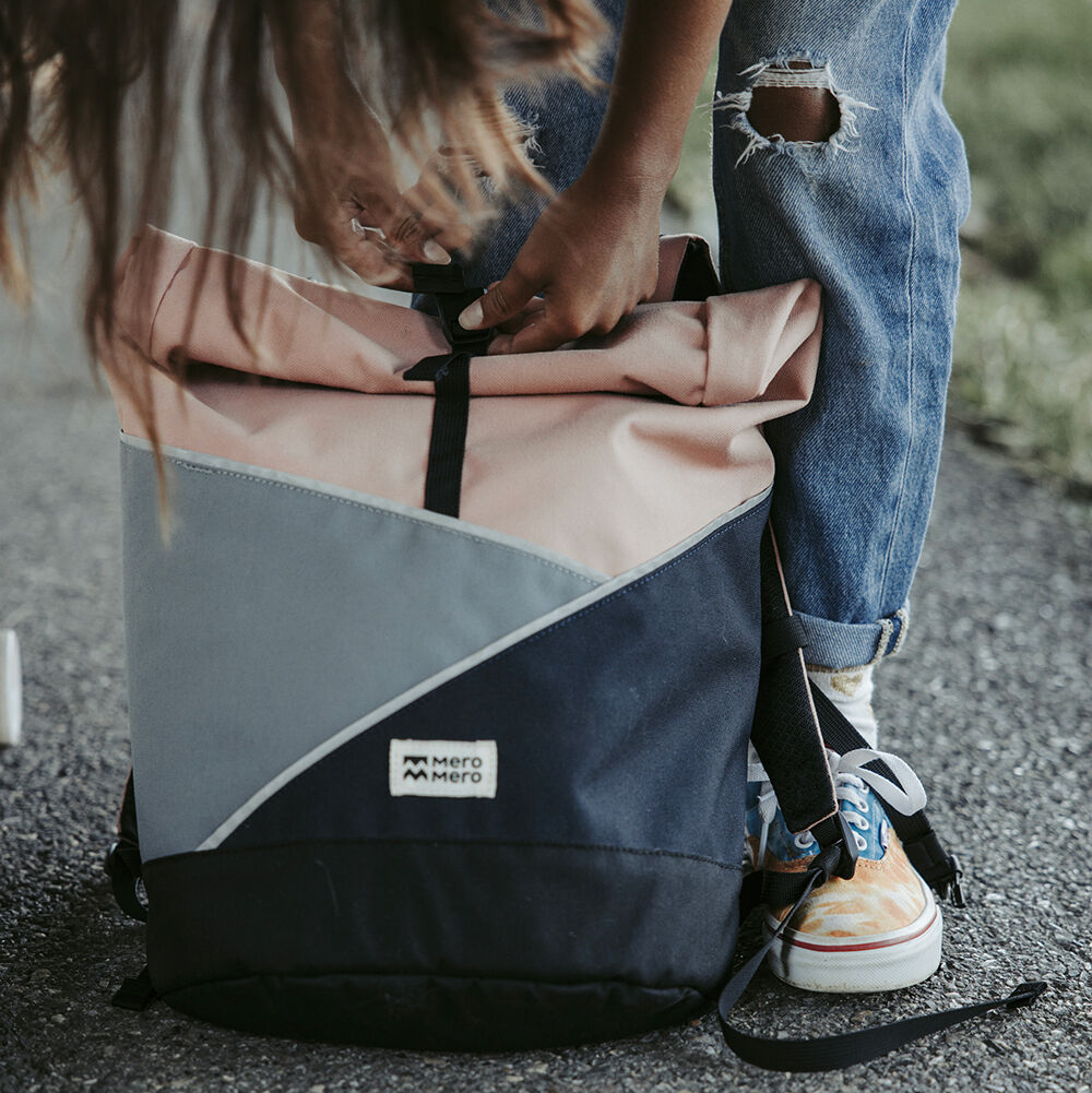 Mensch beugt sich vor, um einen schwarz, hellrosa, weiß und grau gestalteten Rucksack mit Rolltop-Verschluss zu schnüren. Foto im Freien, trägt eine zerrissene Jeans und bunte Sportschuhe.