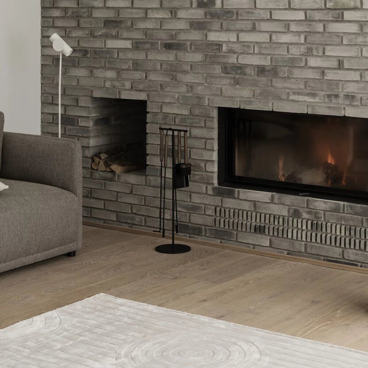 Modern eingerichtetes Wohnzimmer mit grauer Backsteinwand, integriertem Kaminfeuer und Kaminbesteck-Set 'ASHI' aus schwarzem Metall. Daneben ein graues Sofa, ein heller Teppich, Holzkorb mit Brennholz und dezente Dekoration, geeignet für gemütliche Herbst- und Winterabende.