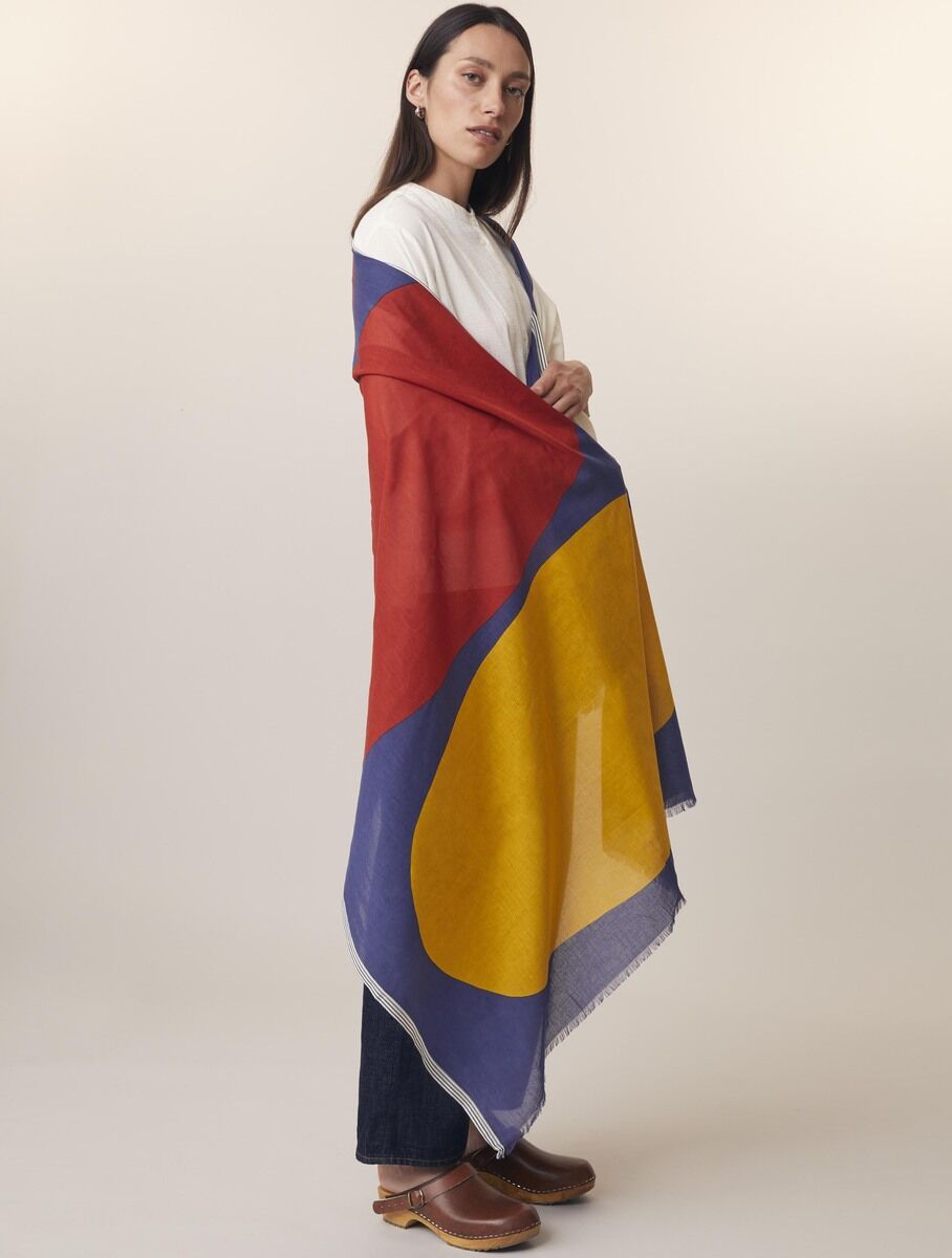 Eine Frau trägt einen Baumwollschal mit bunten Mustern in Rot, Gelb und Blau. Der Schal ist groß und wird über die Schultern drapiert, während die Frau lässig posiert.