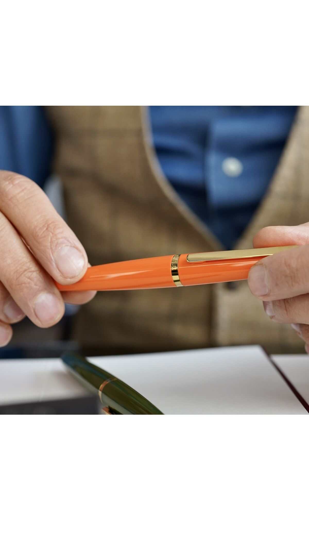 Eine Person hält einen orangefarbenen Füllfederhalter »Design06« mit goldenen Akzenten. Im Hintergrund sind ein blaues Hemd und eine braune Weste sichtbar.