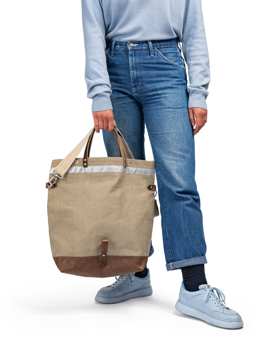 Eine Person in blauer Jeans und hellblauen Sneakers trägt eine beige Fahrrad-Tasche mit braunen und grauen Akzenten.