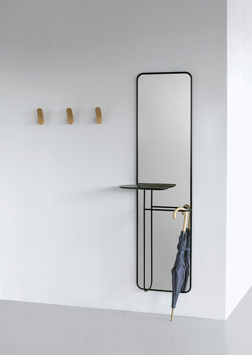 Ein moderner Spiegel namens 'Concierge' hängt an einer weißen Wand. Der Spiegel ist rechteckig und hat einen schwarzen Metallrahmen. Darunter befindet sich eine kleine Ablagefläche aus Glas und an der Seite hängt ein blauer Regenschirm. Weiterhin sind drei Holzhaken an der Wand montiert, die eine minimalistischen Stil unterstreichen.