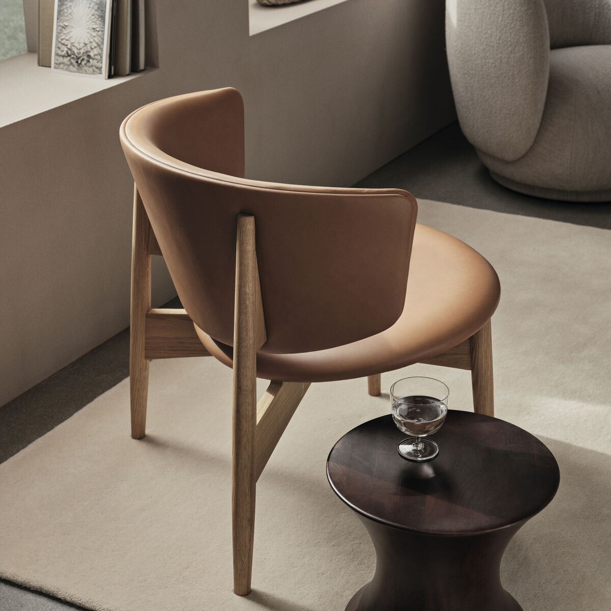 Ein eleganter Herman Lounge Chair mit Holzrahmen und Lederpolsterung, der in einem modernen Wohnzimmer mit minimalistischer Einrichtung steht. Ein kleiner runder Holztisch mit einem Glas darauf ist in der Nähe platziert.