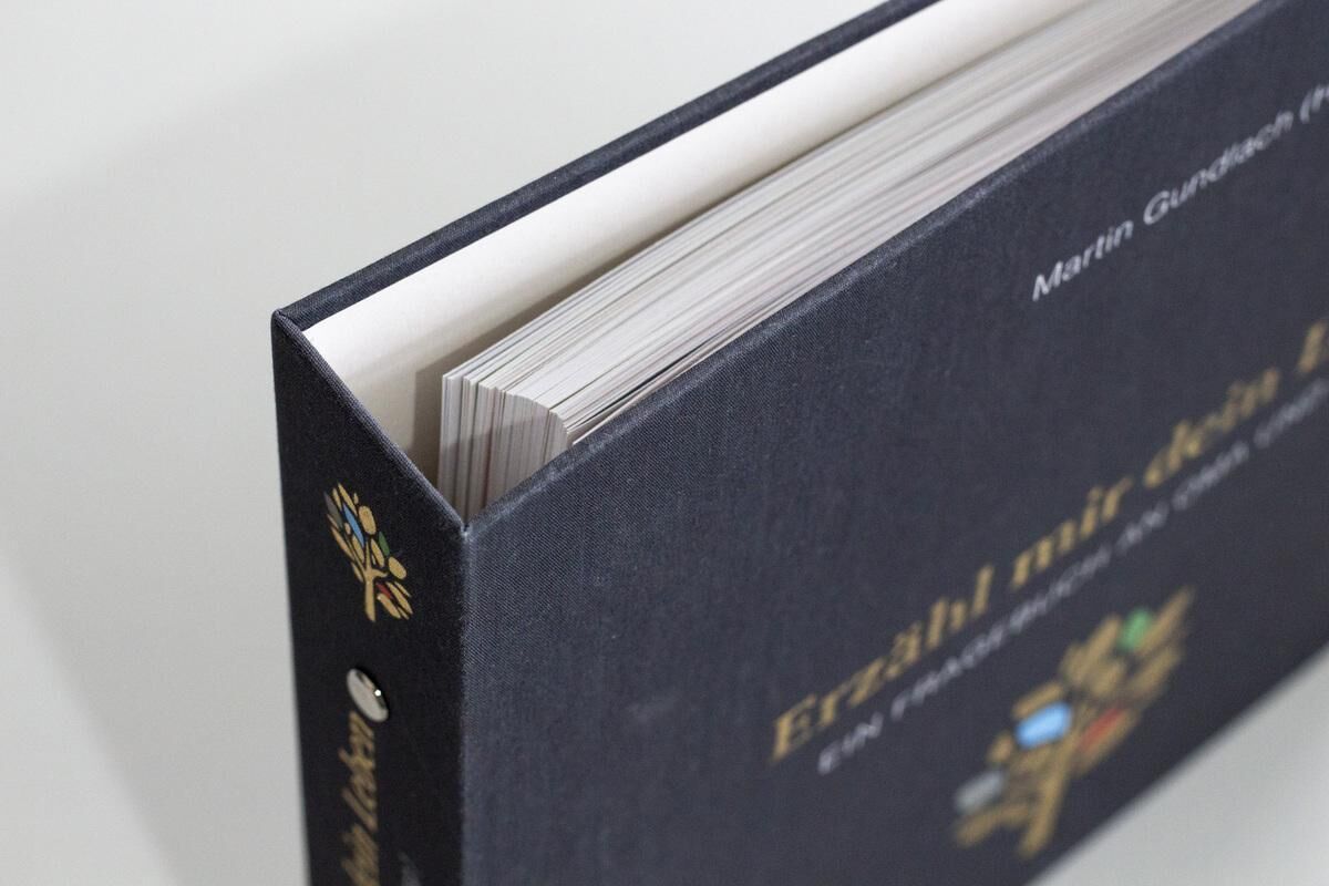 Ein schwarzes Erinnerungsbuch mit dem Titel 'Erzähl mir dein Leben - Leinenausgabe' wird von der Seite gezeigt. Man sieht die dicken Seiten des Buches und einen Teil des Titels in goldenen Buchstaben auf dem Buchdeckel.