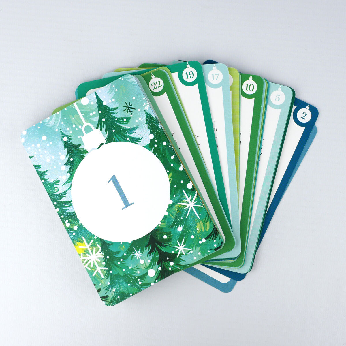 Ein Fächer von Adventskalender-Karten zeigt verschiedene Tage. Die vorderste Karte, nummeriert mit 1, ist mit einem weißen Weihnachtskugel-Motiv und Tannenzweigen auf grünem Hintergrund gestaltet. Die weiteren Karten sind farblich abgestuft und tragen Tageszahlen von 1 bis 22.