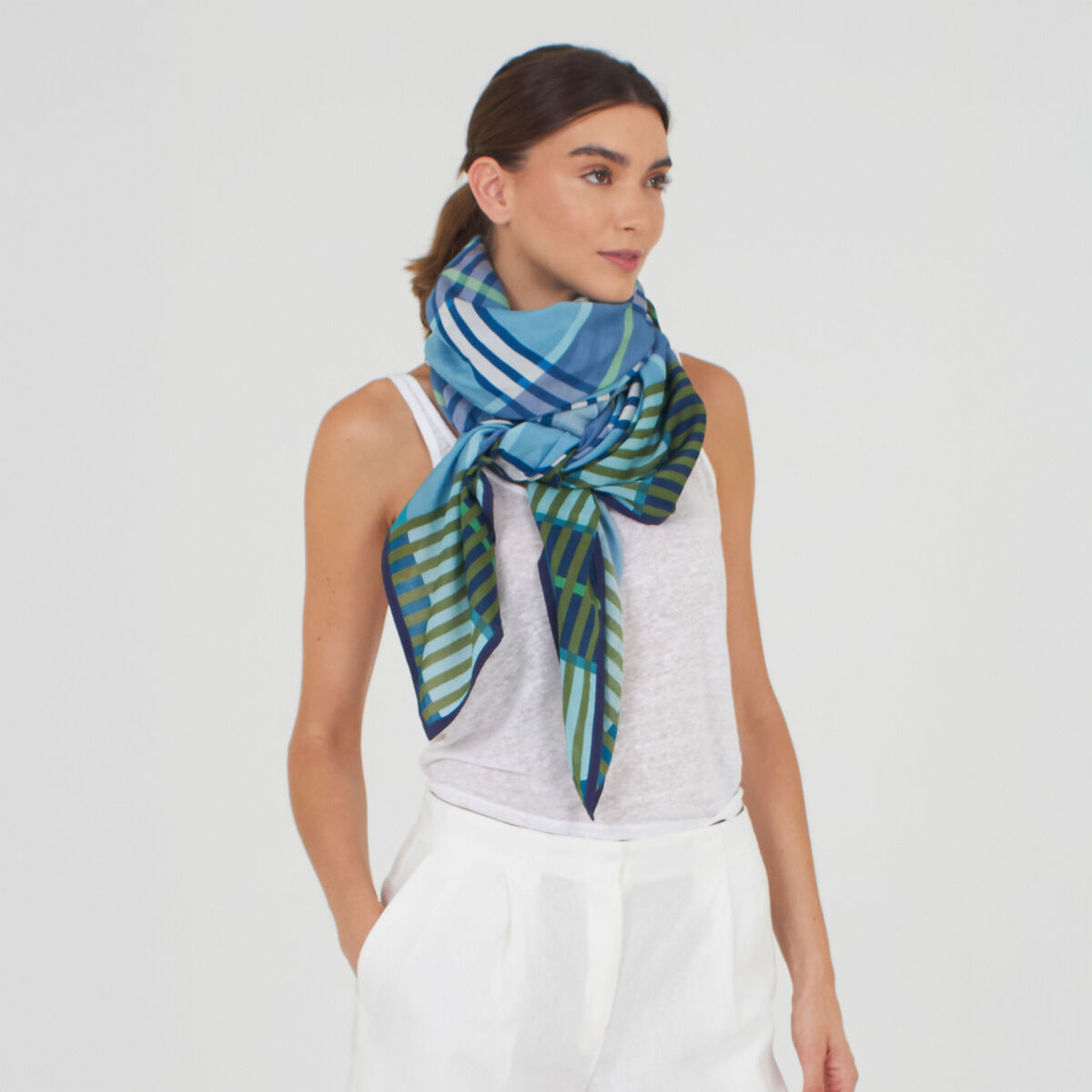 Frau mit schlichter weißer Kleidung trägt den Baumwoll-Seidenschal 'Morgan' Blau von Shalls Barcelona. Der Schal ist großformatig, glänzend und zeigt grafische Muster in Grün-, Blau- und Weißtönen, kunstvoll um den Hals drapiert. Leichte, elegante Optik.