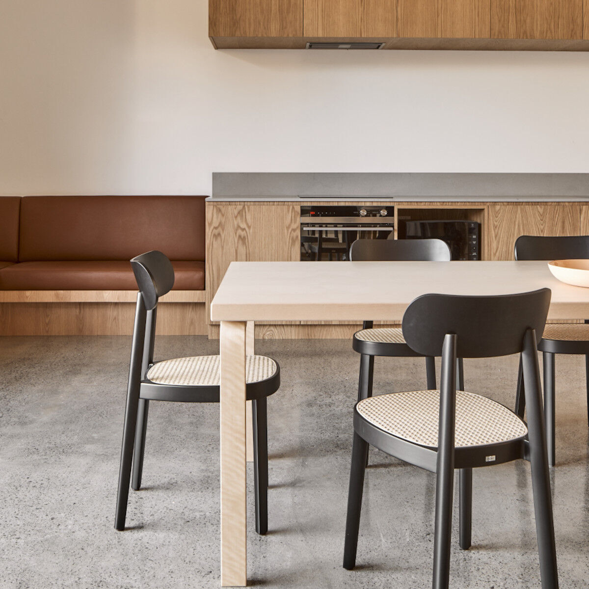 Eine Küche mit einem Holztisch und Thonet Stühlen. Die Stühle haben eine schwarze Struktur und eine Sitzfläche aus beigem Geflecht. Im Hintergrund befindet sich eine Küchenzeile mit Holz- und Metallakzenten.