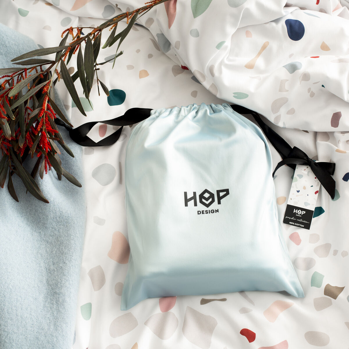 Die Bettwäsche »Terrazzo« zeigt ein modernes, weißes Design mit bunten, terazzoartigen Mustern. Eine hellblaue Stofftasche und ein grauer Zweig mit roten Blütenblatt-Details sind ebenfalls zu sehen.