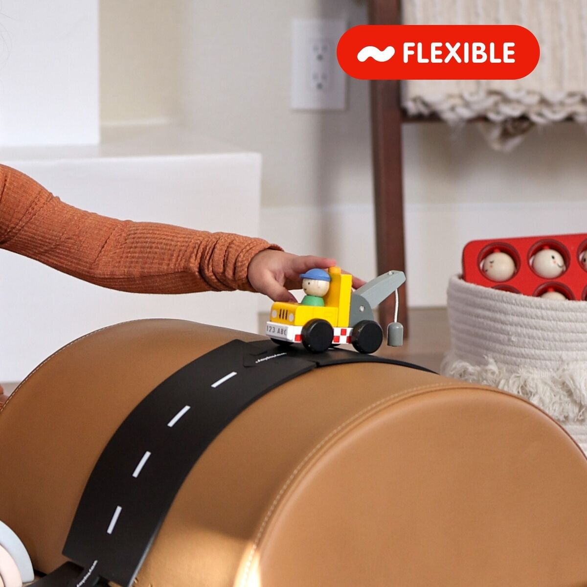Ein kleines Kind spielt in einem Wohnzimmer mit einem flexiblen Straßenbau-Set aus Holz. Das Set umfasst eine schwarze Fahrbahn mit weißen Markierungen, die sich über ein gepolstertes braunes Hindernis schlängelt. Ein Holzauto fährt auf der Straße, weitere Spielzeuge und Figuren sind im Hintergrund sichtbar.
