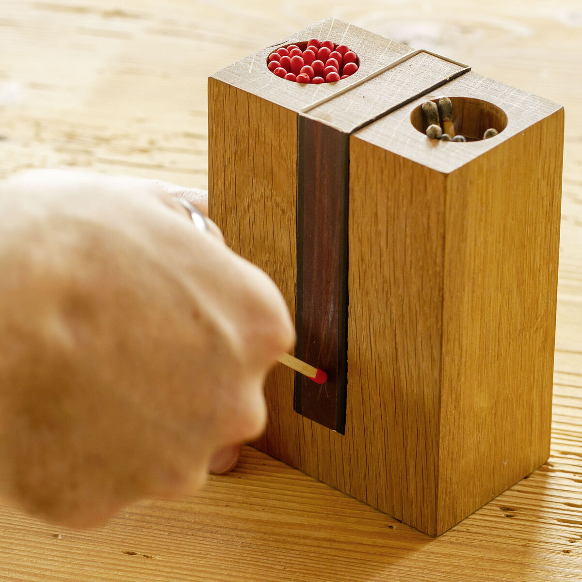 Eine Hand entzündet ein Streichholz an einem Holzblock mit integrierten Streichhölzern und Zündfläche auf einem Holztisch.
