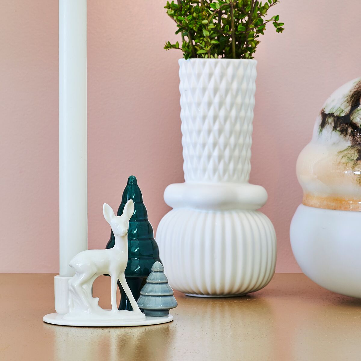 Kerzenhalter aus weißem Porzellan in Form eines stehenden Bambi neben zwei stilisierten Tannenbäumen, platziert auf einem Tisch. Im Hintergrund strukturierte weiße Vasen mit Pflanze und eine weitere Vase mit Farbverlauf auf rosa Wand.