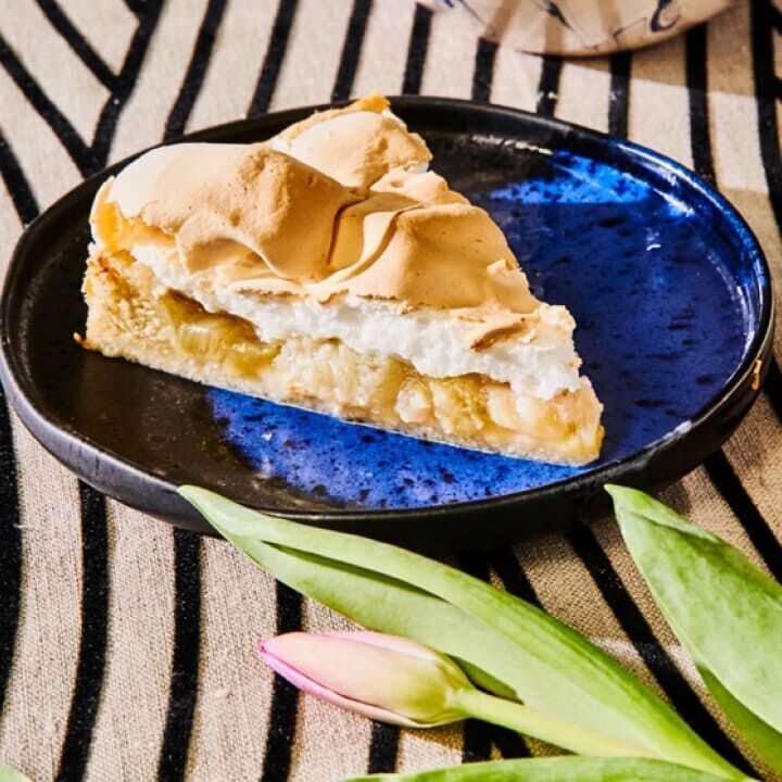 Ein gedeckter Tisch mit einem Teller, auf dem ein Stück Kuchen mit einer Meringue-Decke liegt. Der Teller ist dunkelblau und liegt auf einem gestreiften Tischtuch. Im Vordergrund liegt eine tulpenartige Blume.