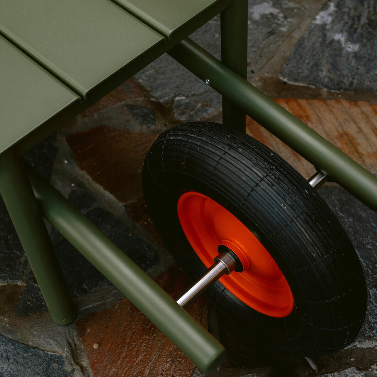 Detailansicht der Schubkarrenbank »Wheelbench« von Weltevree: Olivgrüne Holzlatten und Metallgestänge, darunter ein schwarzes Schubkarrenrad mit orangefarbener Felge. Die Bank steht auf einem nassen, gemusterten Steinboden im Außenbereich.