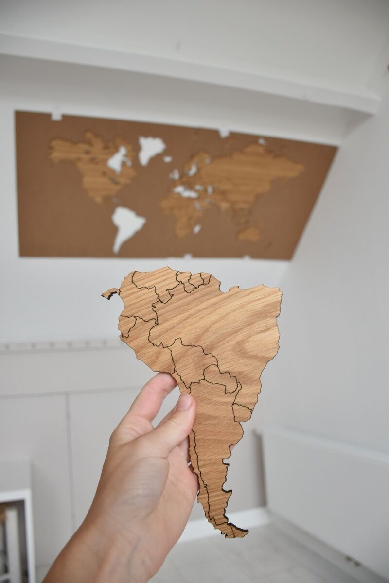 Hand hält ein Stück einer selbstklebenden Weltkarte aus Holz, mit der ganzen Karte im Hintergrund an der Wand angebracht.