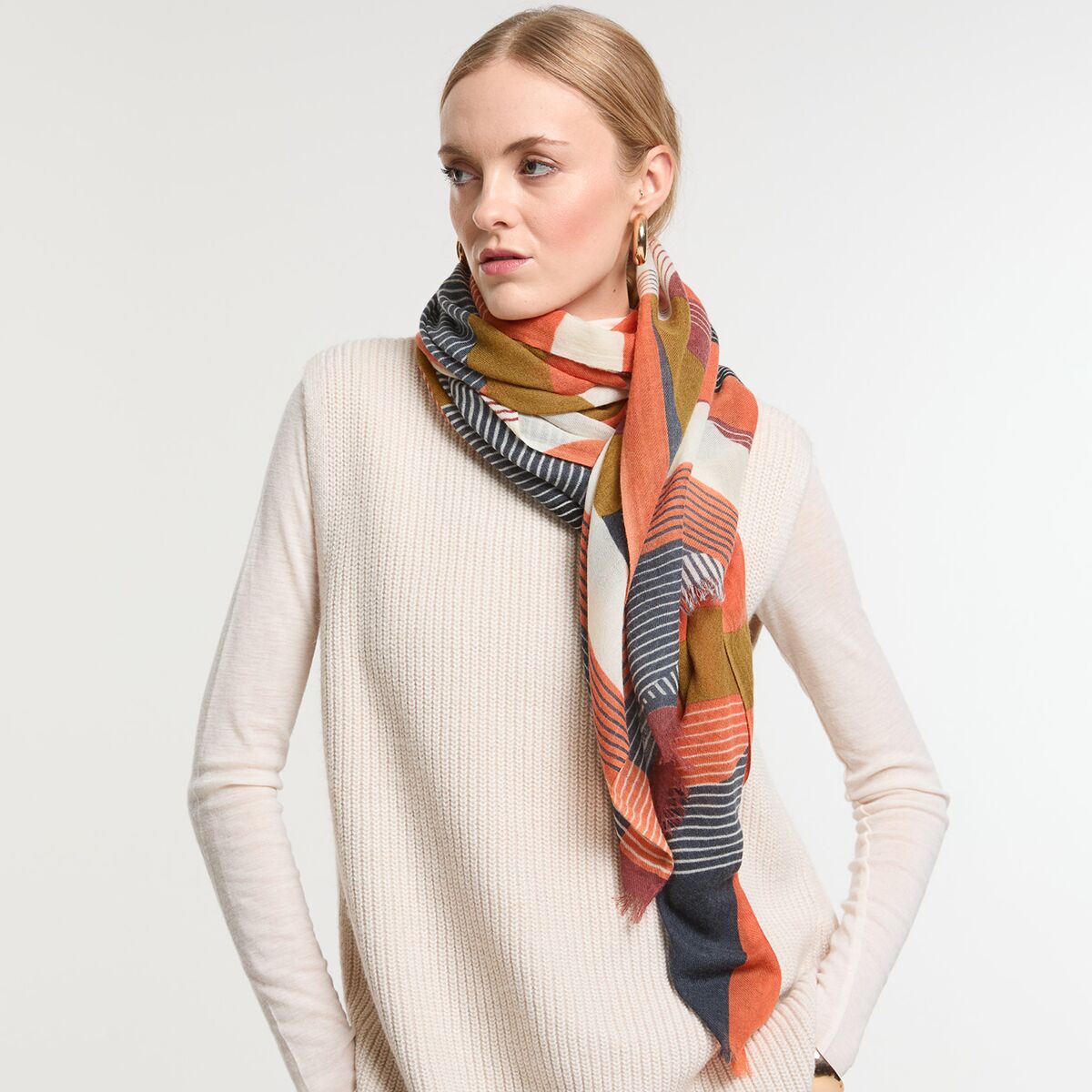 Eine junge Frau trägt einen gestrickten Wollschal in Orange und Rot mit weißen und dunkelblauen Streifen, kombiniert mit einem beigen Pullover. Der Schal wirkt warm und modern, ideal für Herbst und Winter.