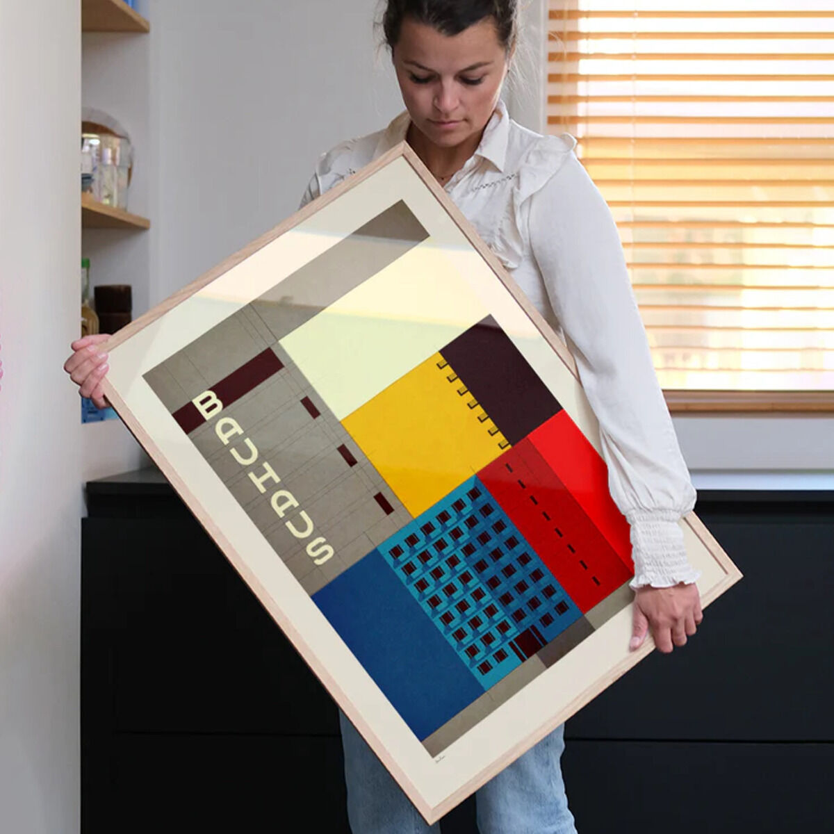 Bauhaus-Poster von Sander Patelski, gerahmt, mit abstrahierten, geometrischen Formen in Rot, Blau, Gelb, Beige, und Schwarz. Eine Person hält das Kunstwerk in einem Interieur mit Holzregal und Fenster.