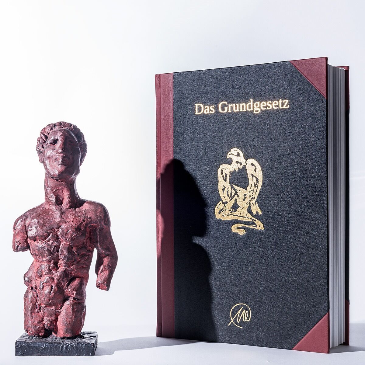 Eine Skulptur und ein Buch mit der Aufschrift „Das Grundgesetz“. Die Skulptur ist dunkelrot, das Buch ist hauptsächlich schwarz mit roten Ecken und einem goldenen Adler auf dem Cover.