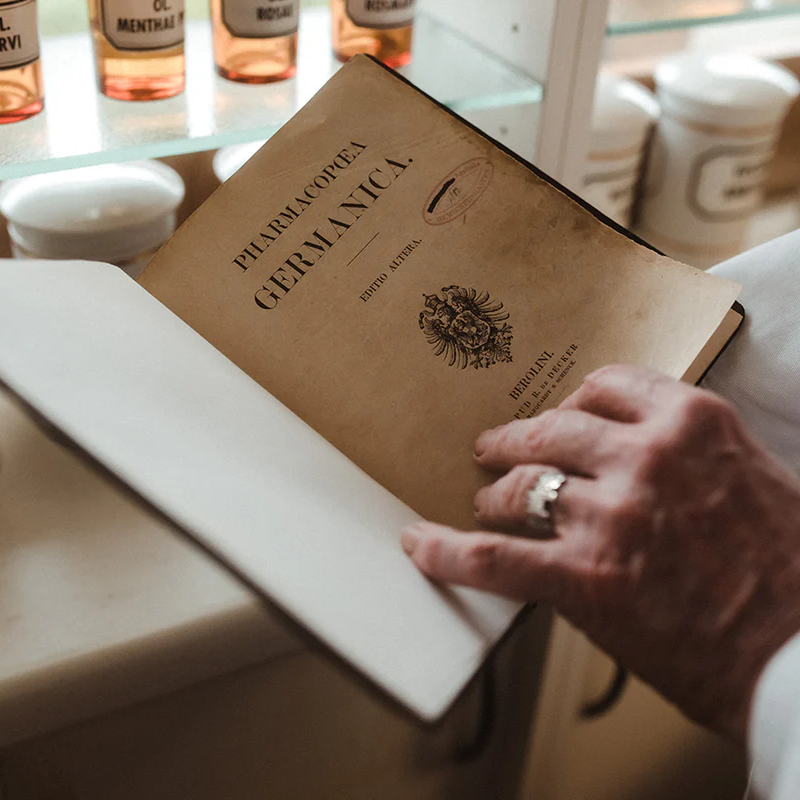 Hand mit Ring blättert in altem Buch 'Pharmacopoea Germanica' auf einem Tisch mit Behältern im Hintergrund