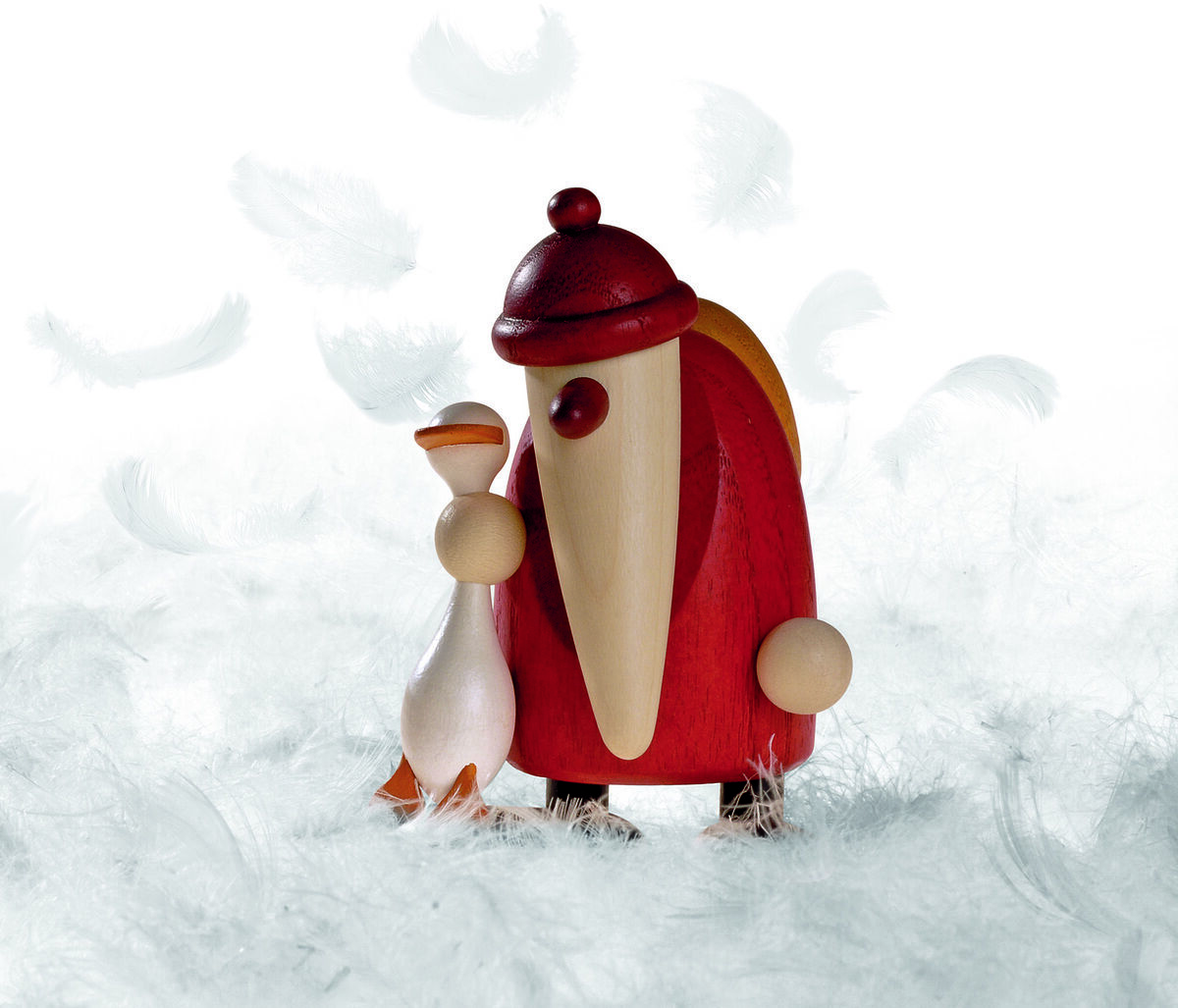 Handgefertigte Holzfigur eines Weihnachtsmanns mit langer Mütze in Rot, gemeinsam mit einer weißen Gans. Die Figuren stehen auf einem weißen, federartigen Untergrund. Die Dekoration vermittelt eine winterlich-festliche Atmosphäre.