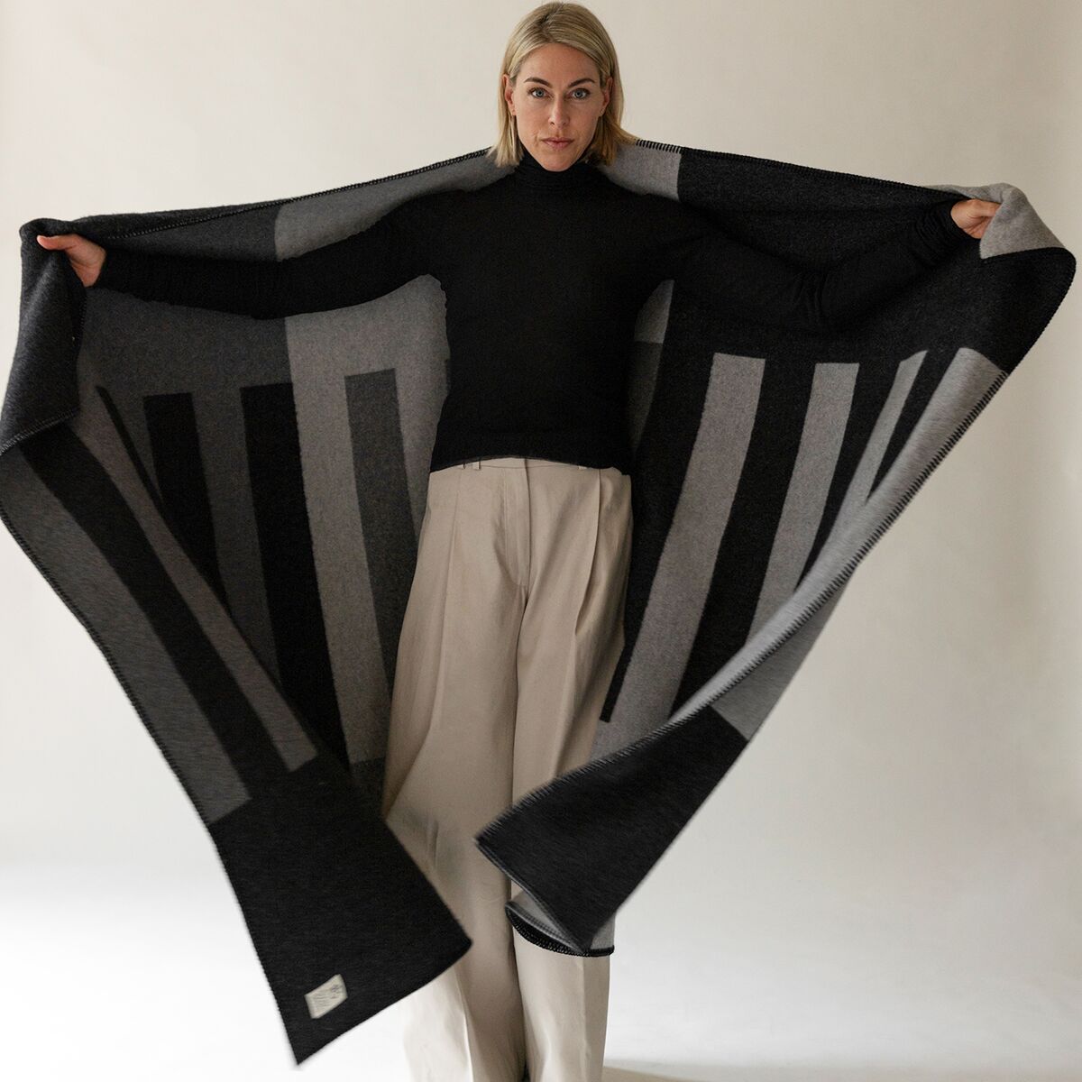 Eine Frau mit blondem Haar trägt eine geöffnete anthrazitfarbene Kaschmirdecke mit grafischen vertikalen Streifen in verschiedenen Grautönen. Die hochwertige Decke ist groß und weich, die feine Webstruktur ist sichtbar, das Etikett unten an der Ecke betont die Markenqualität.