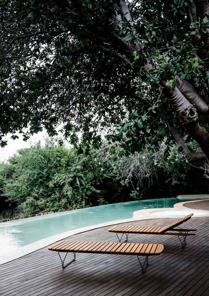 Zwei Holzsonnenliegen stehen auf einer Holzterrasse neben einem Swimmingpool, umgeben von dichter, grüner Vegetation und Bäumen.