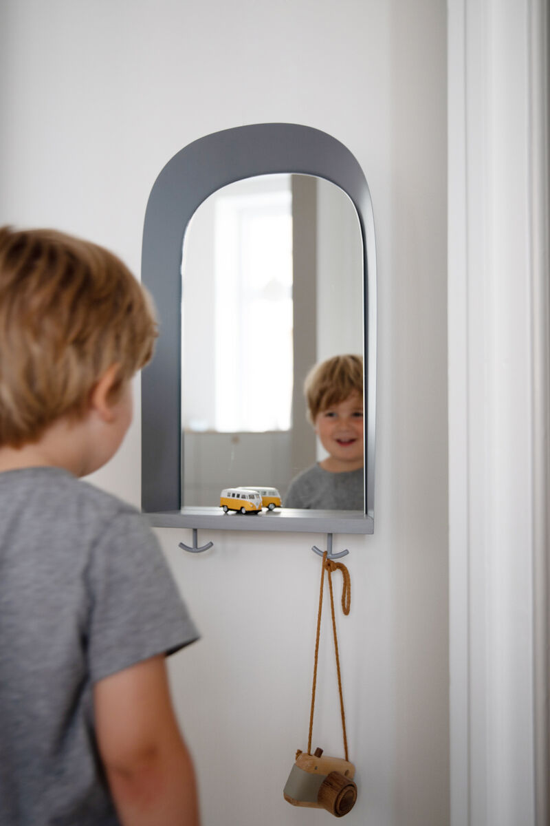 Ein Junge steht vor einem grauen Wandspiegel im Kinderzimmer, unter dem ein kleines Spielzeugauto und eine Holzspielzeugkamera hängen. Der Spiegel hat abgerundete Ecken und der Raum ist in neutralen Tönen gehalten.