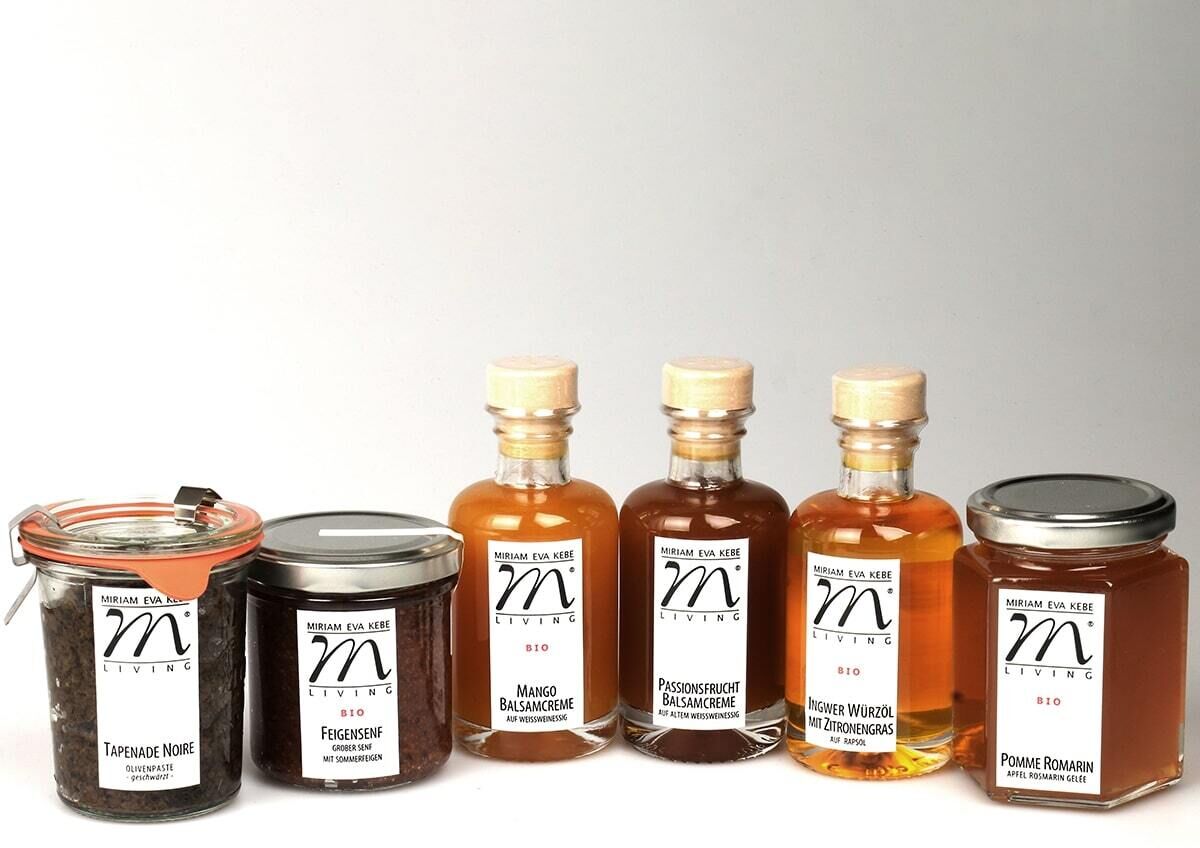 Das Bild zeigt ein Gourmet-Set namens 'ZEIT für Genuss', bestehend aus sechs hochwertigen Produkten in Glasflaschen und Gläsern. Die Produkte umfassen verschiedene Essige, Öle und Marmeladen, die in klaren und bernsteinfarbenen Flaschen präsentiert werden.