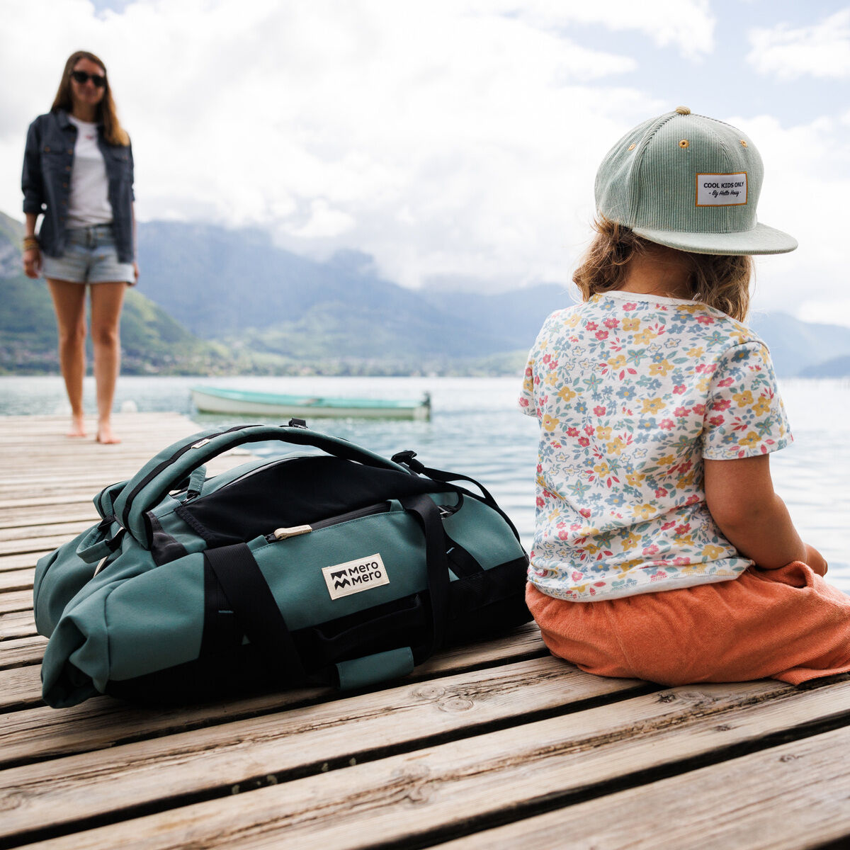 Ein Kind sitzt auf einem Holzsteg am Ufer eines Sees neben einer grünen und schwarzen Mini Smögen Duffle Reisetasche. Im Hintergrund ist eine Frau zu sehen sowie Berge und ein Boot auf dem See.