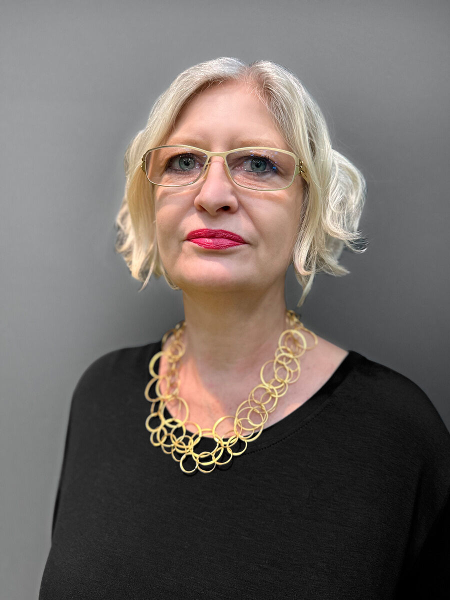 Eine Frau mit blonden Haaren und Brille trägt eine Halskette aus vergoldetem Silber, die aus ineinandergreifenden Ringen besteht. Sie trägt ein schwarzes Oberteil und hat einen neutralen Gesichtsausdruck.
