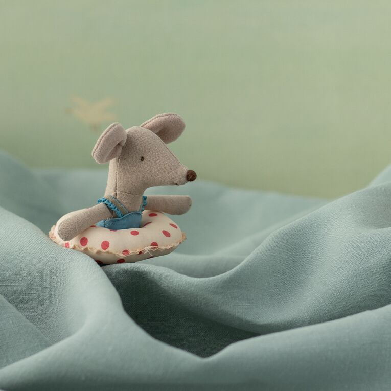Eine kleine, graue Stoffmaus mit blauen Trägern sitzt auf einer bunt gepunkteten Luftmatratze vor einem pastellfarbenen, blauen Hintergrund. Die Szene wirkt verspielt und sommerlich.
