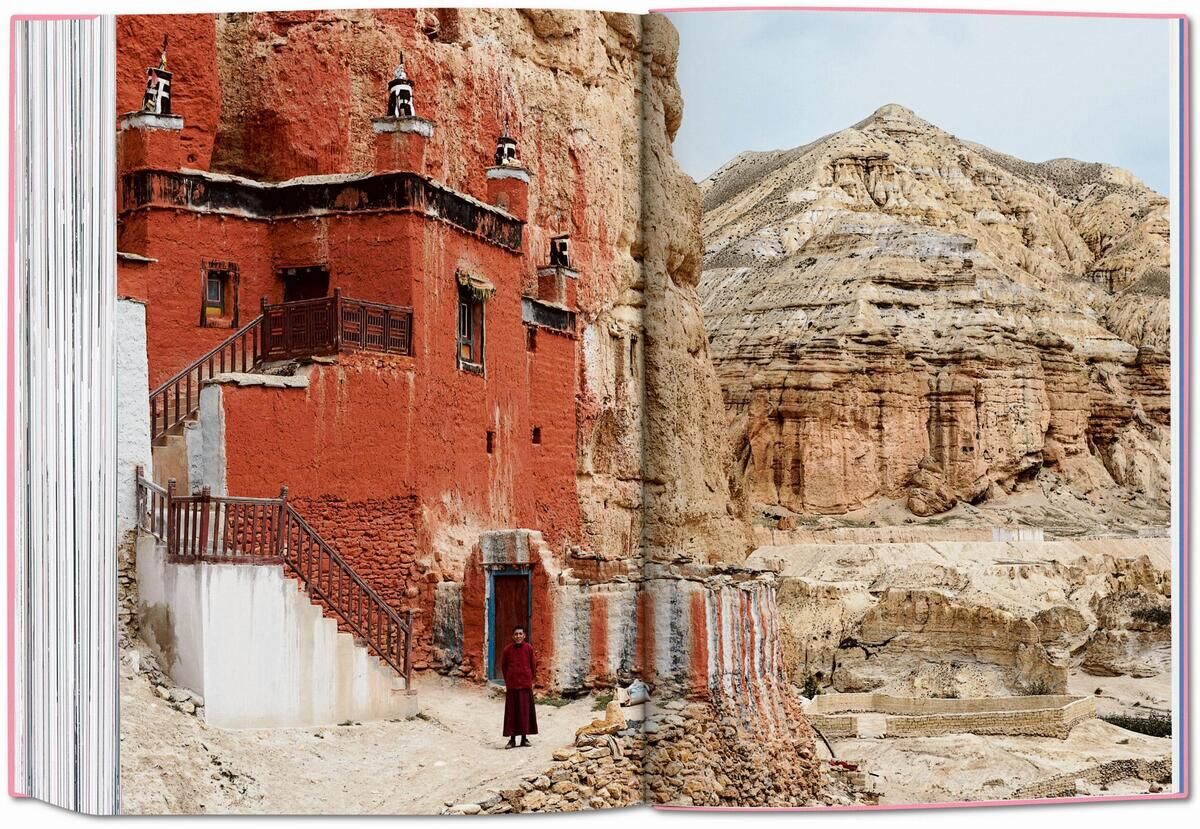 Das Bild zeigt ein aufgeschlagenes Buch mit dem Titel "The New York Times Explorer. 100 Reisen rund um die Welt". Eine der dargestellten Seiten zeigt ein rötliches Gebäude eingebettet in eine felsige, beige Landschaft.