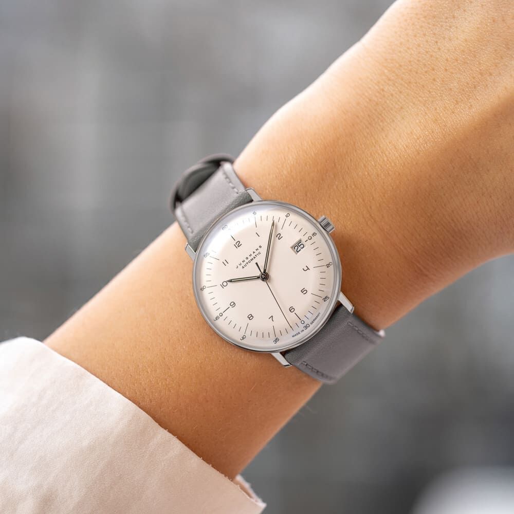 Eine elegante Armbanduhr der ZEIT-Kollektion »Max Bill Kleine Automatic«, getragen an einem Handgelenk. Die Uhr hat ein schlichtes, rundes Zifferblatt in Weiß und ein graues Lederarmband. Sie zeigt ein minimalistisches Design mit klaren Linien.