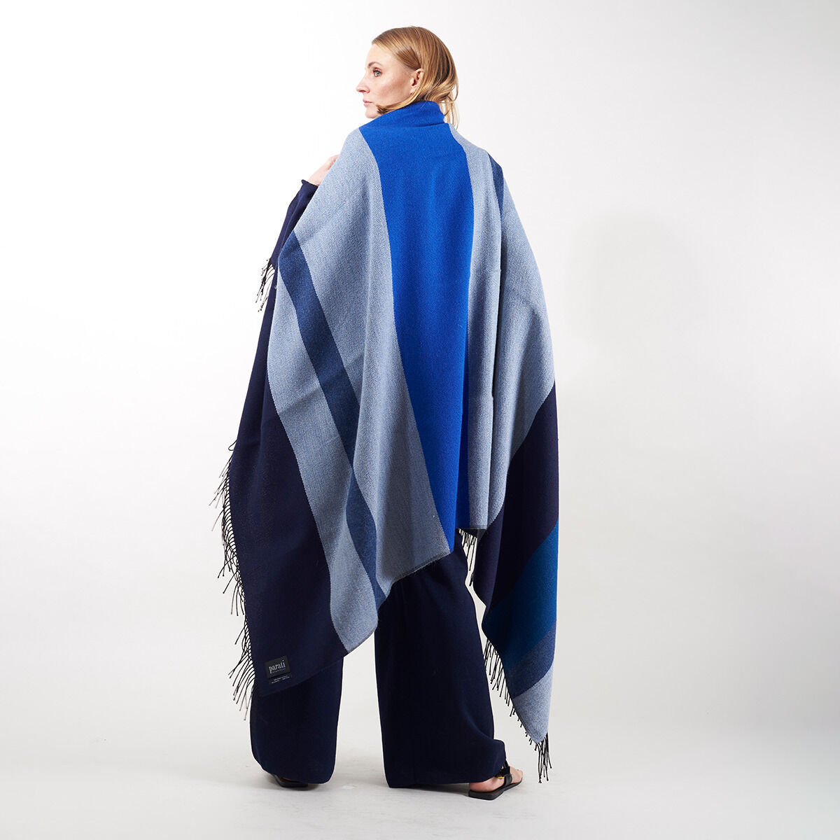 Eine Frau trägt ein Cape aus der ZEIT-Jubiläumsedition »Shades of Blue«. Das Cape zeigt verschiedene Blautöne und hat Fransen an den Enden. Es ist stilvoll und elegant und für den Herbst und Winter geeignet.