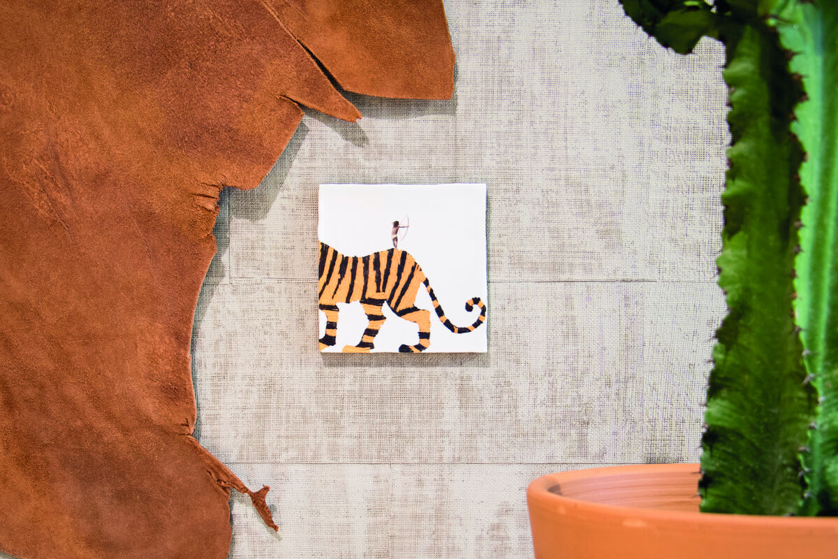 Keramikfliese mit der Abbildung eines Tigers, an der Wand befestigt. Links befindet sich ein Stück braunes Leder, rechts eine grüne Pflanze in einem orangefarbenen Topf.