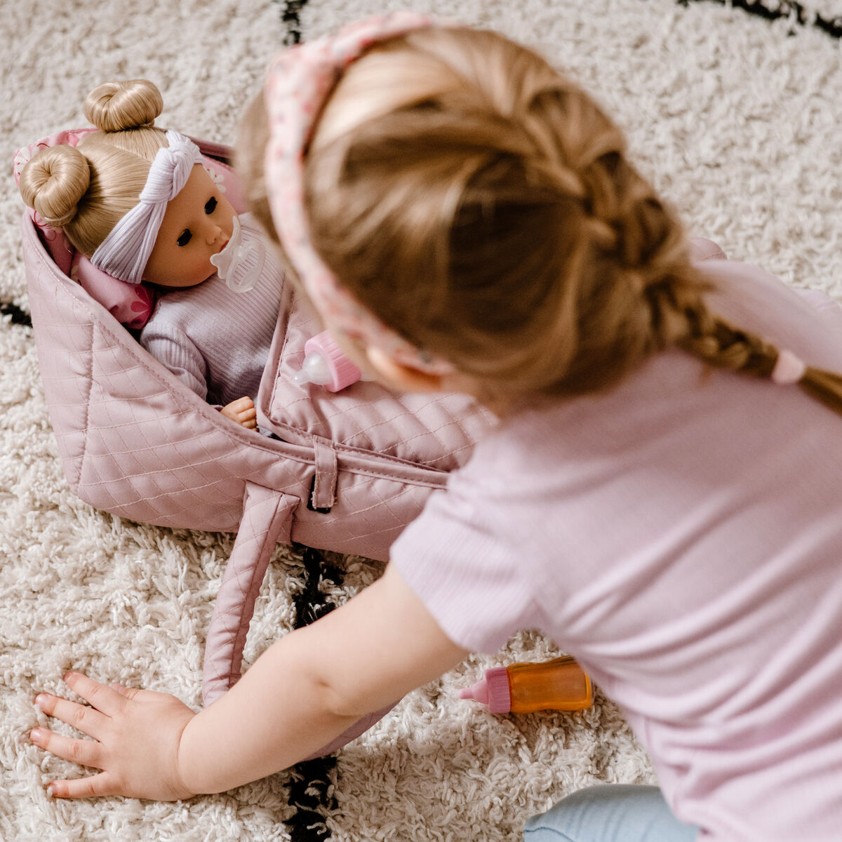 Ein kleines Kind spielt auf einem hellen Teppich mit einer weichen Puppe namens Muffin BliBlaBlume in einer rosa Tragetasche. Die Szene vermittelt Geborgenheit und Freude im Kinderzimmer.