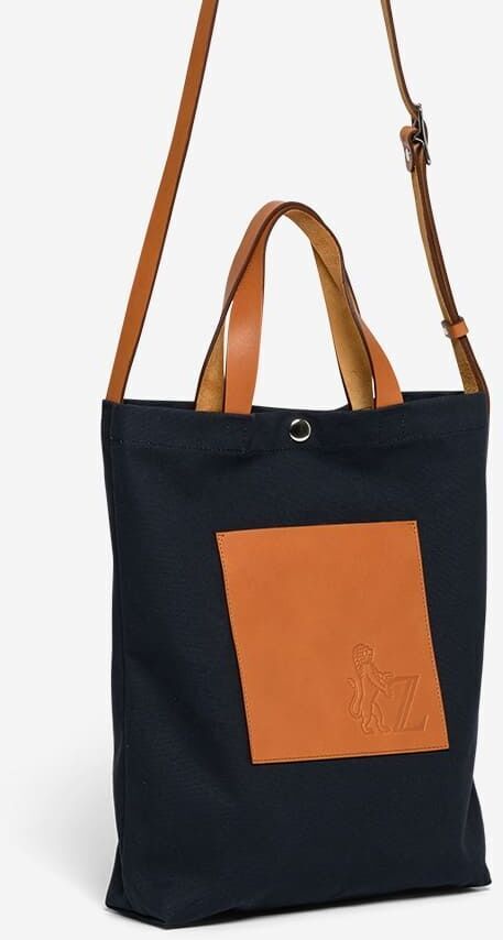Die ZEIT Shop Jubiläums-Shopper Tasche ist eine stilvolle, schwarze Stofftasche mit hellbraunen Lederträgern und einer kleinen, quadratischen Tasche an der Vorderseite.