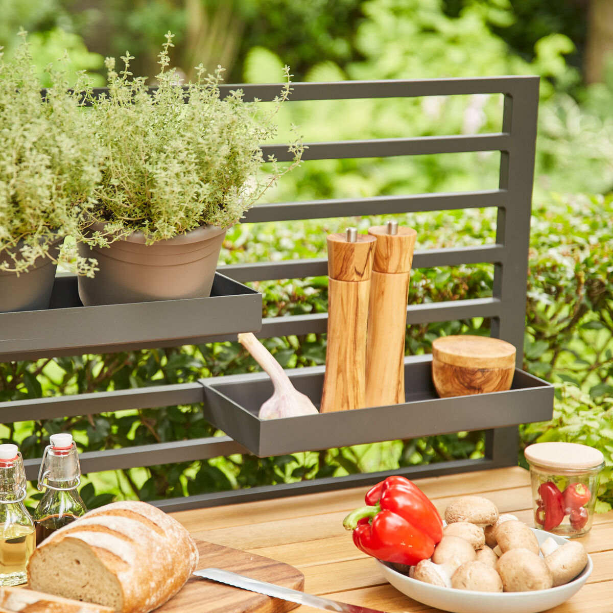 Outdoor-Küche Mittelteil 'Sierra' mit einem Metallgitter, Holztischplatte und verschiedenen Küchenutensilien wie Töpfe mit Kräutern, Paprika, Brot und Holzbesteck, im Hintergrund grüne Sträucher.