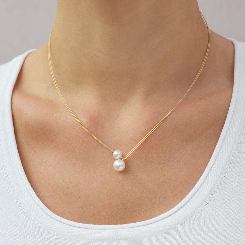 Eine schlanke goldfarbene Halskette mit zwei weißen Perlen, die nebeneinander als Anhänger arrangiert sind. Die Kette liegt elegant am Hals einer Frau, die ein weißes Oberteil trägt. Der Hintergrund ist neutral gehalten.