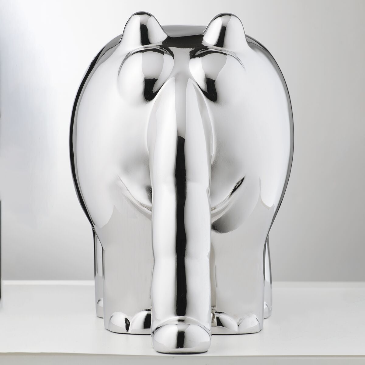 Die Skulptur zeigt einen stilisierten Ottifanten in glänzendem Metallic-Silber. Die Figur steht aufrecht auf einer glatten, weißen Fläche und ist rundlich geformt. Erkennbar sind charakteristische Formen wie der lange Rüssel und die großen Ohren. Das Licht reflektiert die glatte Oberfläche.