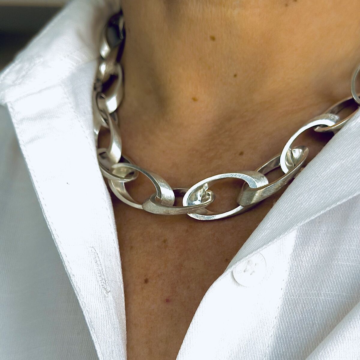 Nahaufnahme eines silbernen Colliers namens "Jackie" um den Hals einer Person, getragen über einem weißen Hemd. Die Kette besteht aus ovalen Gliedern, die elegant und modern wirken.