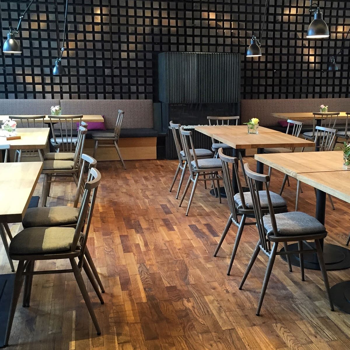 Bild eines modern eingerichteten Essbereichs mit Ercol All Purpose Chairs in grau gebeizt. Die Stühle und Tische sind aus Holz, und die Wand ist ein dunkles Gittermuster. Lampen hängen über den Tischen, auf denen Dekorationen stehen. Der Raum hat einen gemütlichen und modernen Charme.