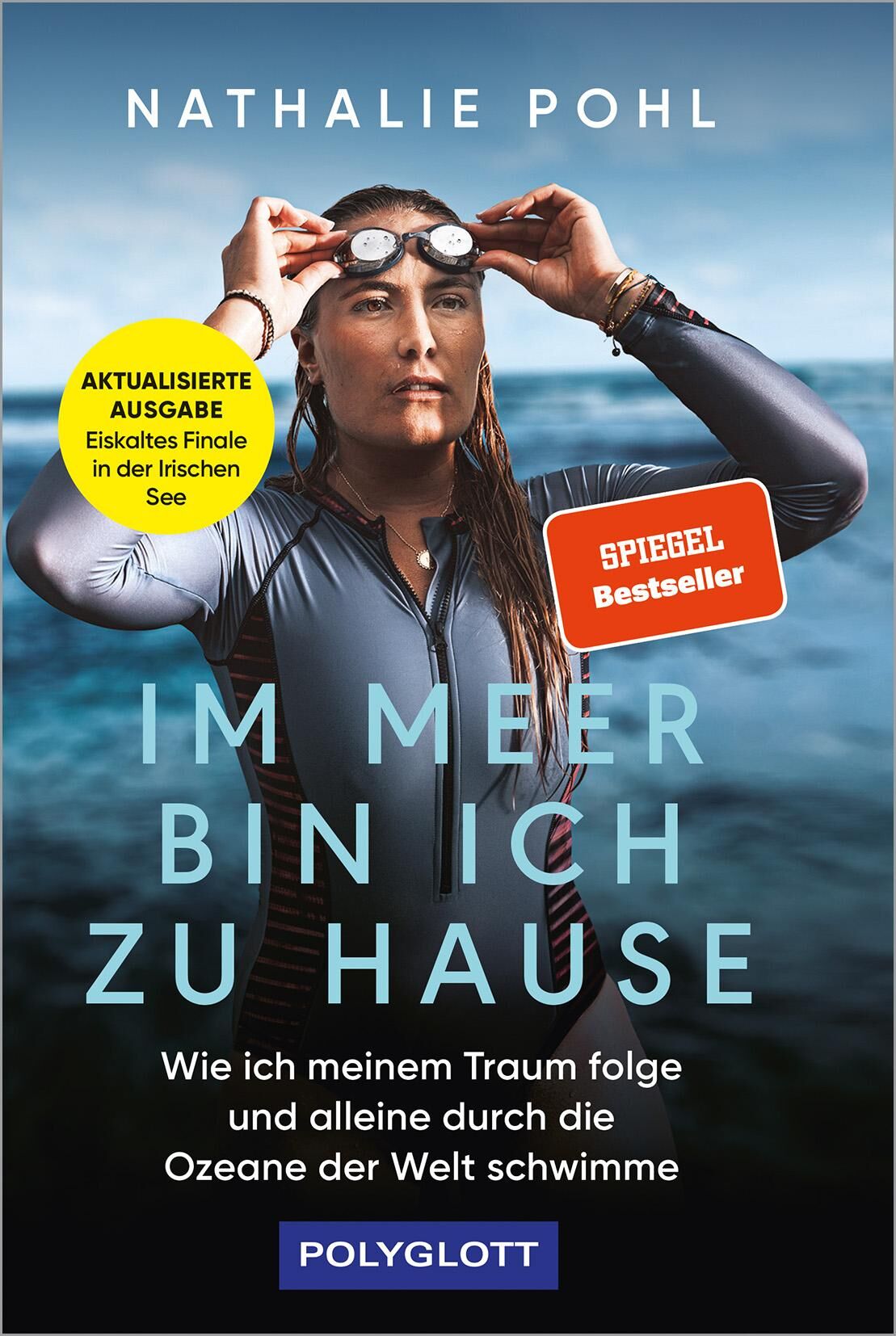 Das Buchcover zeigt Nathalie Pohl, eine Frau in Neoprenanzug und Schwimmbrille am Kopf, die vor einem offenen Meer bei bewölktem Himmel steht. Der Titel 'Im Meer bin ich zu Hause' und der Untertitel stehen im Vordergrund. Die Farben sind hauptsächlich Blau- und Grautöne, unterbrochen von gelben und orangen Akzenten.